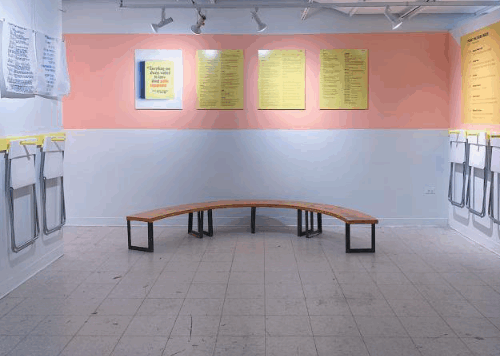 Student work on a gallery wall painted partially pink. There is a bench in the middle of the gallery and folding chairs are hung along the wall for visitors to use.