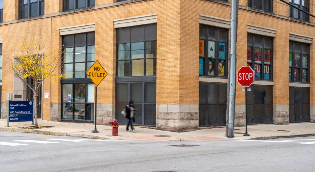 A person is walking down the sidewalk in front of a beige brick building. There is a sign in front of the building which indicates it is UIC's Art and Exhibition Hall located at 400 S Peoria St.