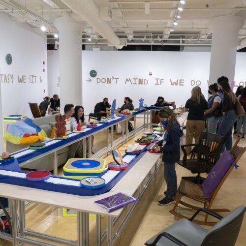 People standing in the Gallery with work on the walls and on a conveyor belt in the center of the room. There are many chairs placed around the conveyor belt.