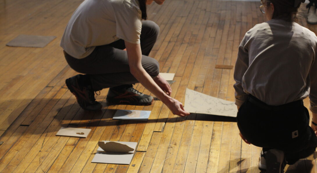 Two students kneeling on a wooden floor. There are small pieces of paper and other objects laid about on the floor. One student is looking at the other who is picking up a larger sheet of paper.