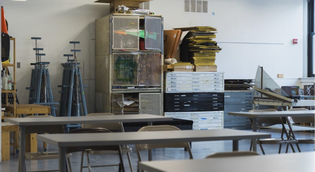 Interior view with tables and metal folding chairs sitting in the foreground. Storage lockers, flat files, and easels line the back wall of the room.
