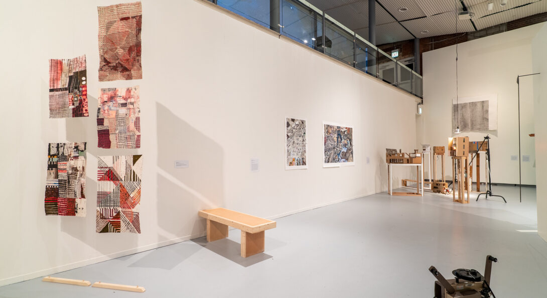 In the foreground on the left hand side is a work by Claire Burke Dain which includes five intricately drawn on pieces of poster-sized paper suspended from the ceiling. The drawings are mostly red and black on off-white and beige paper. Behind the suspended paper, sits a work by Laurel Hauge which is a simple wooden bench. In the background are two works hung on the wall and a number of standing sculptures. A film projector sits in the foreground in the bottom right corner.