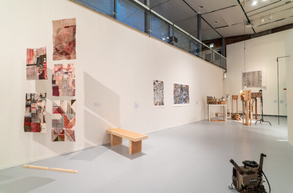 In the foreground on the left hand side is a work by Claire Burke Dain which includes five intricately drawn on pieces of poster-sized paper suspended from the ceiling. The drawings are mostly red and black on off-white and beige paper. Behind the suspended paper, sits a work by Laurel Hauge which is a simple wooden bench. In the background are two works hung on the wall and a number of standing sculptures. A film projector sits in the foreground in the bottom right corner.